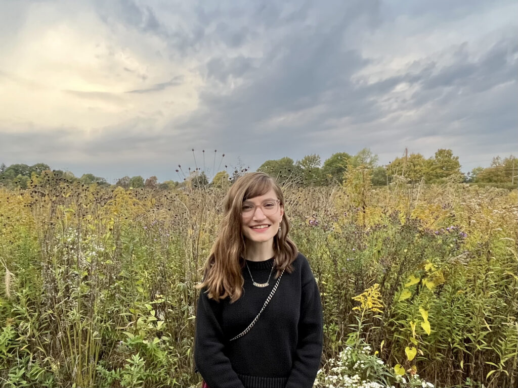 Allison, a white woman with brown hair and glasses, stands in front of a field of yellow, green, and brown autumn flowers